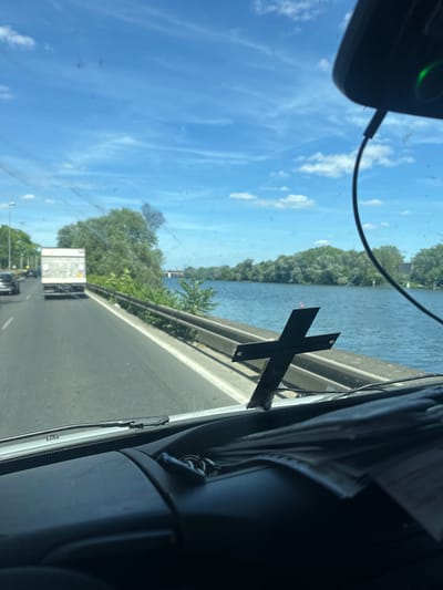 The image, taken from inside a vehicle, shows a view of a highway with a river running parallel to it. The scene appears to be in Argenteuil, France, based on the visible landscape. The highway is occupied by vehicles, including a truck with a white trailer. A distinctive black X-shaped object is attached to the windshield, partially obstructing the view. The sky is blue with scattered clouds. A dashboard camera is visible in the upper left corner of the image. The interior of the vehicle is partially visible on the right side. The focus is on the external environment visible through the windshield. No people are directly visible in the frame.