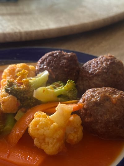 The image displays a close-up of a prepared meal served on a blue-rimmed plate. This scene is situated in Ciudad Victoria, Mexico. The dish prominently features at least three dark brown, spherical items, consistent with meatballs, partially submerged in a reddish-orange sauce. Interspersed among the meat items are cooked vegetables, including orange slices of carrot, green florets of broccoli, and yellowish-white florets of cauliflower. The background is a shallow depth-of-field blur of a light brown surface, likely a tabletop. No individuals or specific actions are depicted.