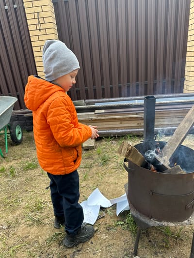 A child wearing an orange jacket and gray hat stands near a metal fire pit in слобода Старая, Russia. Wood and burning material are inside the pit, with smoke rising. Nearby is discarded white paper, and the ground is covered with grass and dirt. A metal fence and a brick structure are visible in the background.