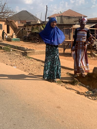 The image depicts an outdoor street scene in Egbedi, Nigeria, under bright daylight with a partially cloudy sky. An asphalt road occupies the foreground, slanting from the lower left to the middle right. Adjacent to the road, two adult females stand on an elevated, uneven dirt and gravel shoulder. The female on the left wears a blue hijab with a scalloped edge, a dark skirt patterned with teal and white motifs, and green flip-flops; her mouth is open as if speaking or smiling. The female on the right wears a multi-colored patterned headwrap and a long, vertically striped dress in shades of brown, white, and red; she holds a white plastic bag in her left hand and a smaller object in her right hand.
In the mid-ground, to the left, an adult male holding a small child's hand walks away from the camera, approaching a building. Behind them, a multi-story building with a dark hip roof and external stairs is visible. Further right in the background are additional buildings, including one with a rusted corrugated metal roof and exposed concrete block walls. Utility poles and overhead lines are present throughout the background. A pile of wood and a two-wheeled vehicle are discernible in the mid-ground. Distinct shadows are cast onto the road and ground, indicating direct sunlight.
