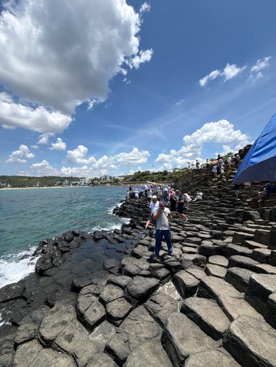 In the city of Phú Lương, Vietnam, a group of tourists gathers on a rocky, volcanic formation adjacent to the sea. The hexagonal basalt columns, a natural geological phenomenon, create a textured, stepped landscape. The sea, a vibrant teal, meets the shore with gentle waves. In the distance, a town with white buildings and lush green hills is visible. The sky above is a brilliant blue, dotted with fluffy white clouds. Several individuals are scattered across the rocks, some taking photos, others conversing. One man in the foreground, wearing a grey t-shirt and blue pants, is facing away from the camera. A large blue tarp, possibly an umbrella or canopy, is set up on the rocks to the right of the frame, offering shade. The overall scene captures a popular tourist destination characterized by its unique natural beauty and the presence of visitors.