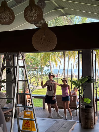 In Pekutatan, Indonesia, a man wearing a white tank top with the number 30 and dark shorts, and a woman in a pink tank top and denim skirt, both wearing hats, stand on a wooden deck. The man is making a peace sign with his left hand and pointing with his right. The woman is pointing to her right. Behind them, palm trees and the ocean are visible through an open doorway. A stepladder is positioned to the left of the couple, with a yellow caution sign at its base that reads "CAUTION WET FLOOR" and "HATI-HATI LICIN." Several large, woven pendant lamps hang from the ceiling above.