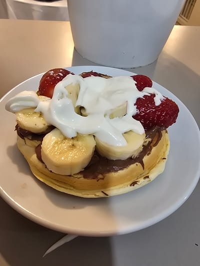 A close-up, high-angle shot features a dessert plate, likely a pancake or crepe, richly topped with fruit and cream, set in an indoor environment. The main subject is a light brown, folded pancake or crepe, generously spread with a dark brown substance, presumably chocolate or Nutella. On top of this, numerous slices of ripe banana and several whole, glistening red strawberries are arranged. A substantial amount of thick, white cream, possibly whipped cream or yogurt, cascades over the fruit and chocolate, creating a visually appealing contrast.

The food is served on a simple, round white ceramic plate. In the immediate background to the left, a large, off-white appliance or piece of furniture is visible, showing some dark smudges near its base. Further left and slightly upwards, a metallic vent or grill can be discerned. The rest of the background consists of muted, light gray and beige surfaces, suggesting a kitchen or dining area. There are no people visible, and no specific activity is taking place beyond the presentation of the food. The lighting appears to be artificial, casting a warm glow in parts of the background, but the time of day is not discernible. No text is visible in the image. The scene is likely set in a domestic interior in La Florida, Chile.