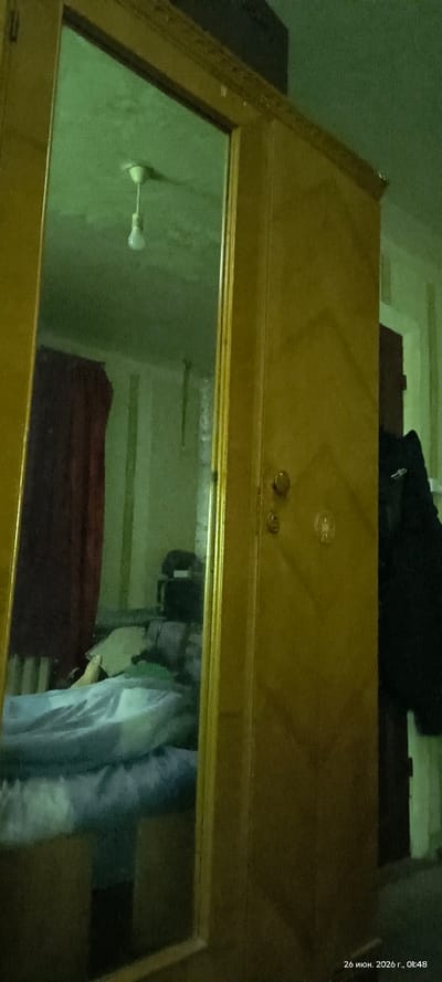The image shows a reflection in a large wooden wardrobe mirror. In the reflection, a person's legs are visible on a bed covered with blue bedding. The room appears to be dimly lit with a green tint. A red curtain is visible to the left of the bed. The wardrobe itself is made of light-colored wood and has a diamond pattern on its door. There is a round doorknob and a keyhole visible on the wardrobe door. A black coat is hanging on the right side of the wardrobe. The bottom right corner of the image contains a timestamp: "26 июня. 2026 г., 01:48". This timestamp suggests the image was taken in Russia on June 26, 2026, at 1:48 AM.