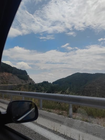 This is a photograph taken from inside a vehicle, looking out towards a mountainous landscape under a partly cloudy sky. The foreground features a car's side mirror and the edge of a road with a guardrail. The road curves along the side of a lush green mountain, which is terraced in some areas with exposed rock and vegetation. In the distance, other green hills are visible. The sky is a bright blue with scattered white clouds. There are no people or animals visible in the image. The scene appears to be a scenic route, possibly in a rural or mountainous area. The location is not identifiable as Komotini, Greece based on the visual information alone.