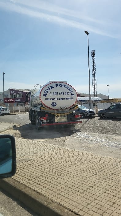 The scene depicts an industrial area within the Polígon Industrial De Calvià, Spain, under a bright, partly cloudy blue sky. A large white tanker truck, viewed from the rear, occupies the center-right of the image. The truck's cylindrical tank features the text "AGUA POTABLE" (Potable Water) in blue, a phone number "608 638 097", and "TRANSPORTE DE AGUA A DOMICILIO" (Water delivery service to homes). The vehicle has a red rear bumper, visible taillights, and mud flaps, with a partially visible Spanish license plate.
The ground directly behind and around the truck appears wet and reflects light, indicating standing water or a recent wetting event on a paved surface of cracked concrete or asphalt. To the right of the truck, two passenger vehicles, a dark sedan and a grey SUV, are parked or stationary. In the background on the left, a multi-story commercial or industrial building with red trim and numerous windows is visible. On the right, a tall, black lattice-style telecommunications tower stands alongside a street light. The immediate foreground shows a section of light-colored, square-patterned pedestrian paving and the dark blue housing of a vehicle's side mirror, from which the photograph was taken. No individuals are visible.