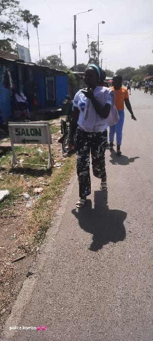 The image depicts a daytime street scene in Homa Bay, Kenya, characterized by clear skies and direct sunlight.
In the foreground, a female subject walks along a paved road, positioned slightly right of the frame's center. She wears a light blue headband, a white short-sleeved top with a graphic, camouflage-patterned trousers, and light-colored open-toed sandals. A white fabric item is draped over her left shoulder, and her right hand holds a dark object near her mouth, suggesting communication. Her distinct shadow extends onto the pavement.
To her immediate right and slightly behind, another individual walks, wearing an orange t-shirt and light blue trousers. Further down the road, additional pedestrians are visible, receding into the distance.
On the left side of the frame, a grassy verge with scattered debris and dry vegetation parallels the road. On this verge, a white wooden A-frame sign with "SALON" in green capital letters and a left-pointing green arrow stands upright. Behind the sign, a dark-colored two-wheeled vehicle, possibly a motorcycle, is partially visible. Further left, a row of simple, single-story structures with corrugated roofs lines the street. One prominent structure is painted blue, featuring an open doorway revealing at least two seated individuals inside. Various items, possibly clothing, hang outside these structures.
The background features tall utility poles equipped with streetlights, and scattered trees, including prominent palm trees, under a bright, cloudless sky.