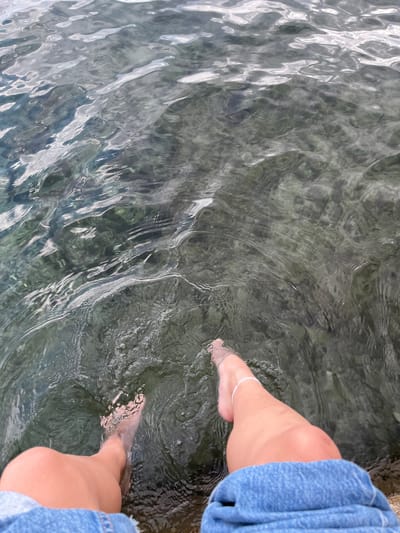 Two bare feet are submerged in a body of water. The water appears clear, and the bottom is visible, consisting of rocks and pebbles. A person is seated, as suggested by the denim-clad legs on the right edge of the frame. An anklet adorns the left ankle. The scene is likely in Grad Opatija, Croatia, given the presence of clear water.