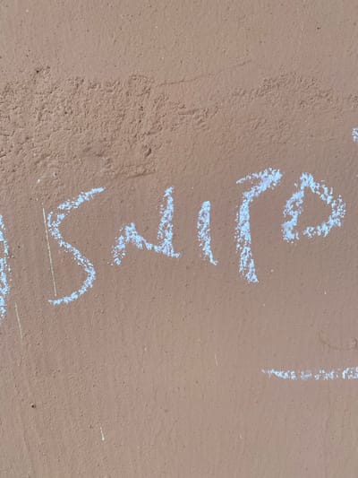 The image shows chalk graffiti on a brown, rough-textured wall. The graffiti appears to be a word or phrase written in white chalk. The letters are somewhat fragmented and uneven, suggesting they were drawn with haste or on a rough surface. A single faint yellow vertical line is visible to the left of the white chalk writing. The background is a uniform brown wall, likely an exterior surface. The image is an abstract close-up of the writing and wall, with no discernible context for its location in Dumnga, Nigeria.