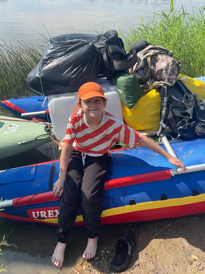 A young person wearing an orange baseball cap and a striped t-shirt sits on a blue and red inflatable raft, facing the camera with a smile. Their legs are bare and dangling in the water. The raft is loaded with various bags, including a large black trash bag, backpacks, and a yellow waterproof bag. The raft is positioned at the edge of a body of water, with tall green grass growing along the bank. The ground is muddy. In the background, the water surface is visible under a clear sky. The scene appears to be a natural outdoor setting, possibly a riverbank or lake shore. The location is identified as Nezhinka, Russia.