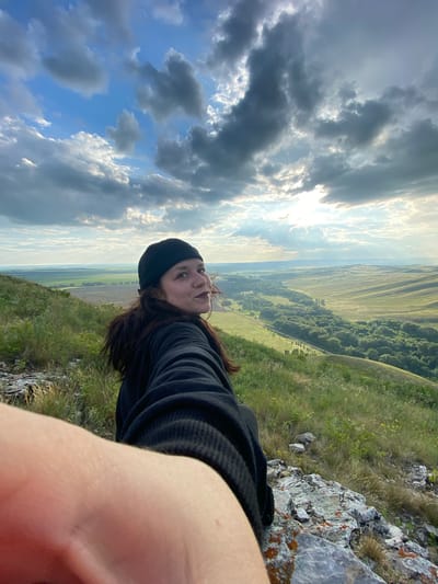A young woman wearing a black beanie and a black hoodie is sitting on a rocky, grassy incline, looking back over her shoulder at the camera. Her right arm is extended forward, obscuring part of the frame. The background reveals a vast, undulating landscape with green fields and a winding treeline. The sky is partly cloudy with the sun peeking through, casting rays of light. The location is the vicinity of Petrovskoye, Russia.