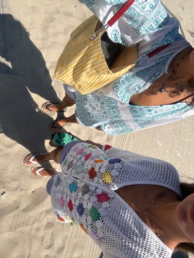 Two individuals are observed from an elevated perspective, standing on light-colored sand, indicative of a beach or dune environment in the city of Caminha, Portugal. Strong, elongated shadows cast to the left suggest bright sunlight.
The individual in the foreground, positioned to the right, wears a white, open-knit crochet cover-up adorned with multicolored floral motifs in shades of pink, blue, green, orange, yellow, and gray. A delicate gold-toned body chain with small reflective elements is visible on their torso. Their legs are brown-skinned, and they wear white flip-flops. The chin and mouth area of this individual, with dark hair, are partially visible on the far right edge of the frame.
The second individual, positioned in the background to the left, wears a white cover-up with a teal patterned design. They carry a yellow and white striped canvas tote bag with red handles, slung over their left shoulder. The brand "Bimba y Lola" is subtly visible on the bag. A dark beaded necklace with a circular pendant rests on their chest. Their legs are also brown-skinned, and they wear black flip-flops. A rolled-up green object, possibly a towel or mat, is situated near their feet.