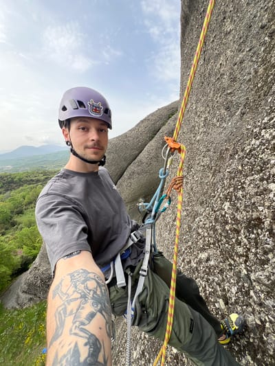 This vertical, eye-level shot captures a male rock climber positioned against a textured, grey rock face. The climber, a young adult with short dark hair and stubble, wears a light purple climbing helmet adorned with a colorful cartoon monster sticker, a grey t-shirt, and olive green climbing pants that display the "SIMOND" logo. His left arm is heavily tattooed with intricate black and grey designs.
The climber is equipped with a grey climbing harness, a yellow and red patterned climbing rope that passes through an orange and silver belay device attached to the harness via carabiners. Additional carabiners and slings are visible on his harness. A black and yellow climbing shoe is seen on his right foot, pressed against the rock.
The perspective suggests the climber is either actively climbing, belaying, or at a high anchor point on the rock face. The immediate foreground is dominated by the rough, imposing rock. Beyond the climber, a vast and verdant landscape unfolds, featuring lush green trees and distant mountains under a partly cloudy sky. The bright daylight and clear visibility indicate favorable outdoor conditions. The distinctive, massive rock formations and mountainous backdrop strongly suggest the location is Kastraki, Greece, specifically the Meteora region. The climber maintains a neutral, focused expression as he looks towards the camera.