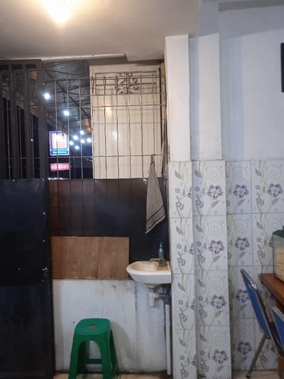 The scene is a small, white-walled bathroom with tiled walls and a partially visible door. The walls are decorated with a floral pattern. A small, white sink sits on a counter with a towel hanging above it. A green plastic stool is in front of the sink. An unidentifiable object is on the counter near the sink. There is a metal grate to the left of the sink with a sign visible behind it. The scene is dimly lit and no one is present. It is located in Kecamatan Kartasura, Indonesia at -7.558 latitude and 110.777 longitude.