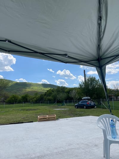 The image depicts a partly obscured view from under a tent. In the background, a landscape with a green hill, trees, a blue sky, and white clouds is visible. A blue car is parked near a fence. A white chair and a bottle of water are on the right side of the image. A wooden pallet is on the ground. The location is likely Klenovik, Bulgaria.