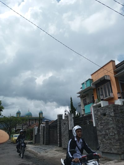 The image displays a street scene in Cibadak, Indonesia, under an overcast sky with dense grey clouds. In the foreground, right of center, a male individual wearing a white full-face helmet, a black jacket with white arm stripes, and a light-colored shirt is operating a white motorcycle. Further down the street on the left, a second male individual, wearing a dark helmet and dark attire, rides a black motorcycle. A yellow car is partially visible behind the second motorcyclist, near foliage.

Architectural features dominate the background. On the left, a large mosque with multiple teal-colored domes and minarets is visible behind trees, with mountains indistinctly in the distance. On the right, a multi-story building features an orange upper section and a teal mid-section with large windows. This structure is fronted by a fence constructed from stacked grey stone with dark metal railings. Another, closer stone-clad fence with a decorative light fixture is visible on the right foreground. Overhead, a dark power line stretches diagonally across the sky. The road surface is asphalt.