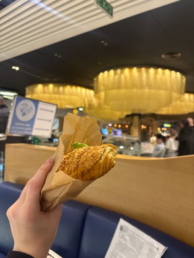 A person's right hand, with fair skin and manicured fingernails, is centrally holding a sesame-seed-covered, croissant-shaped pastry partially enclosed in a brown paper bag. A green leaf is visible within the pastry. The individual is situated in an indoor commercial space within Bubuieci, Moldova. The background features a dark ceiling with multiple large, circular, gold-toned chandeliers providing illumination. An illuminated green "EXIT" sign is visible on the upper left ceiling area. In the midground, a blue upholstered bench seat and a light wooden partition are present, with a white document resting on the seat to the right. To the left, a blue sign with white pictograms is partially visible. The distant background is blurred, showing several indistinct figures of people standing and sitting, suggesting a public dining or café environment with various other light sources and commercial displays.