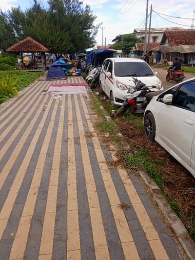 The image shows a paved walkway with alternating black and tan rectangular tiles, located in Pameungpeuk, Indonesia, at latitude: -7.668, longitude: 107.694. The walkway is lined with vegetation on both sides. To the right, there are two parked white cars and three motorcycles. One person, dressed in a blue shirt, is visible walking away from the camera. The image captures a general view of the scene, focused on the paved walkway.  There is a blue tent on the left side of the photo.