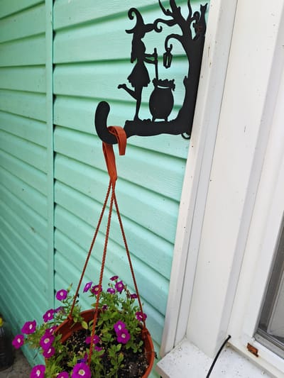 A hanging basket filled with purple petunias is suspended from a black metal hook attached to a light green, horizontally paneled wall. The hook is shaped like a silhouette of a witch with a cauldron and a cat. To the right of the wall is a white window frame. The scene appears to be outdoors, possibly on a porch or balcony. The location is the city of Unarokovo, Russia.