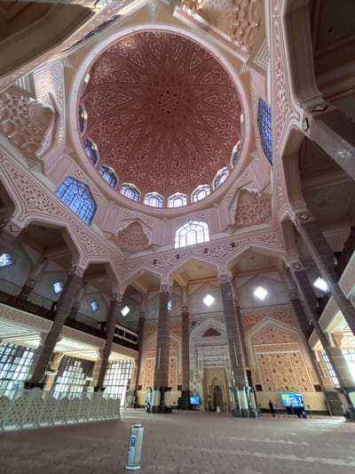 The image shows the interior of the Putra Mosque in Putrajaya, Malaysia, looking upwards towards the grand dome. The architecture features intricate pink and white decorative patterns on the dome and surrounding arches, supported by tall, dark columns. Blue stained-glass windows are visible along the upper walls. The floor is covered in a light-colored carpet, and a low, decorative barrier runs along the left side of the image. Several people are present, mostly standing or walking in the distance. Digital screens are mounted on some of the pillars. Sunlight streams in through large windows on the left side of the mosque.