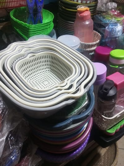 A stack of nested, square-shaped colanders dominates the image. The colanders are varying shades of gray and white. Beneath them is a stack of colorful, similar shaped dishes. To the right are various plastic containers and bottles with colorful lids, and a basket holding a water bottle. A stack of green plastic dishes are near the top left of the image. A tall stack of dark plates is situated behind the green dishes.