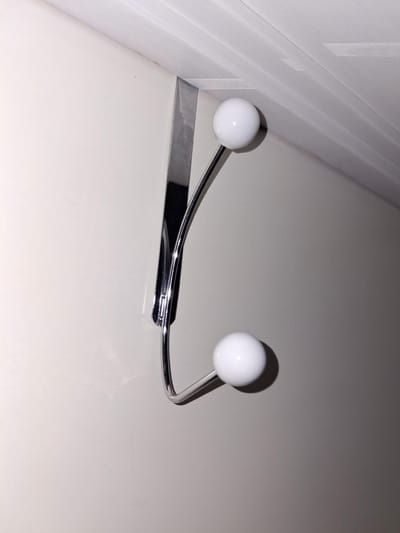 A chrome over-the-door hook with two white ball-shaped ends hangs on a plain, light-colored wall, near a white ceiling. The hook is positioned vertically, with the top hook section resting on the top edge of the door and the curved section extending downwards. The lighting creates shadows on the wall. The image does not depict any people or activities, and the location in Åsgårdstrand, Norway, is not visually represented in the image itself.