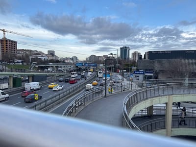 The image depicts an urban landscape in Gungoren, Turkey, under an overcast sky characterized by extensive grey cloud cover. In the immediate foreground on the right, a multi-level, curved concrete pedestrian overpass with silver metal railings extends. A single individual, wearing a light-colored coat, is observed walking downwards on a lower section of this overpass.

The midground features a multi-lane highway with moderate to heavy traffic flow. Vehicles include numerous private cars, several yellow taxi cabs, white delivery vans, and multiple passenger buses. A dark-colored sedan with a yellow and black diamond-shaped sticker on its rear passenger door is visible in one lane. To the left of the highway, a yellow construction crane stands beside a multi-story building frame under active construction. A group of approximately five individuals is visible standing on an elevated green-roofed structure spanning an intersecting road.

The background on the right is dominated by a large "Mercedes-Benz Otomol" dealership building, identifiable by its dark, textured facade and prominent signage. A "VOLVO" sign is also visible below the Mercedes-Benz sign. Various other high-rise commercial and residential buildings populate the distant horizon. Overhead road signs displaying text such as "e-haciz yok" and "e-devlet Kapınız" are present above the highway. The scene captures a bustling urban environment with ongoing development and active transportation infrastructure.