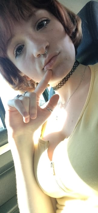 The image features an individual with reddish-brown hair and bangs, possessing light-colored eyes and a silver septum piercing, facing the viewer. They are wearing a light-colored, front-zippered garment, a black choker necklace, and a thin silver chain. The individual's right hand is raised, with the index finger positioned against their lower lip or chin. Multiple rings are visible on the fingers of the right hand, including a silver band on the thumb. The background suggests an indoor environment, possibly a vehicle interior, with blurred elements and strong natural light originating from the right side of the frame, causing significant highlights and overexposure on the lower right portion of the subject's body and clothing. The specific location within the city of Goričani, Montenegro, is not ascertainable from the visual content of the image.
