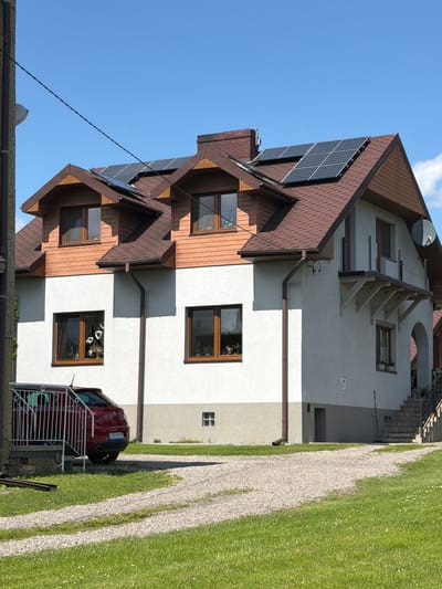 The image shows a two-story house with a brown shingle roof and white walls, situated in Strzyżowice, Poland. The house features multiple windows with brown frames, and several solar panels are installed on the roof. A red car is parked in front of the house. The foreground consists of a gravel driveway and a green lawn with scattered white flowers. The sky is clear and blue.