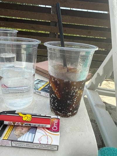 The image depicts a close-up, eye-level view of a table outdoors, likely in Manolsko Konare, Bulgaria, with a wooden fence in the background. On the table, there are two clear plastic cups, one filled with a dark beverage and ice, and the other with clear liquid. A black straw is inserted into the cup with the dark beverage. To the right of the cups, a white plastic lounger chair is partially visible. In the foreground, a pack of cigarettes, labeled "Omé," lies next to a lighter that features a yellow cartoon hand graphic on a red background. A folded pink wallet is also visible between the cups. The table surface appears to be a light-colored, textured material.