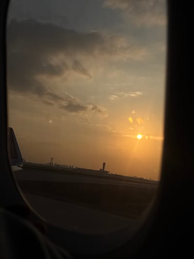The image displays an exterior view from an aircraft window at sunset, captured at Ben Gurion Airport (TLV) in Yehud Monosson, Israel. The scene is dominated by a setting sun in the middle-right, casting an intense orange and yellow glow across the sky, accompanied by distinct lens flares. Overcast and scattered clouds, ranging from dark gray to lighter tones, are visible above the horizon. Below the horizon line, dark airport tarmac or runway surfaces are prominent. A tall, cylindrical air traffic control tower is silhouetted against the sunset in the mid-right background. Beyond the tower, a distant cityscape or industrial area with multiple structures and at least one crane is discernible. In the lower-left foreground, a portion of the aircraft's vertical stabilizer, featuring a purple accent or logo, and a segment of its wing are visible, framed by the dark, rounded window. No individuals are present in the image.