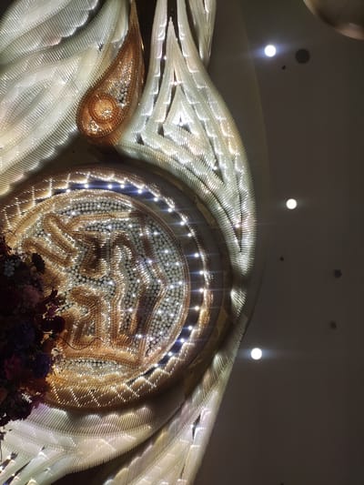 An ornate, crystal-like chandelier hangs from a ceiling in a dimly lit interior space. The chandelier is composed of many translucent, cylindrical elements arranged in a swirling, petal-like pattern, with some elements appearing golden or amber. Embedded within the chandelier are numerous small, bright lights, creating a dazzling effect. In the lower left corner, a cluster of dark purple and pink flowers is visible, possibly part of a floral arrangement integrated with the chandelier or hanging nearby. The ceiling itself is a neutral, muted color, with several circular, recessed lights visible in the upper portion of the image.

The chandelier is located at the Urumqi Grand Bazaar, a large shopping complex in Urumqi, Xinjiang Uyghur Autonomous Region, China.