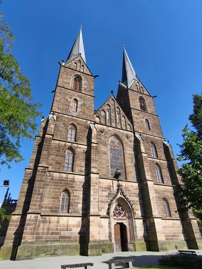 This is a low-angle, full-shot photograph of a large, Gothic-style stone church with two prominent spires, set against a clear, bright blue sky. The church is constructed of large, rough-hewn brown stones, with architectural details such as arched windows and decorative stonework visible. Sunlight casts strong shadows on the facade of the building, indicating it is likely midday or early afternoon on a sunny day. Lush green trees frame the left and right sides of the church, with some branches extending into the frame from the left. In the foreground, there are two modern-looking benches and a paved area, suggesting a public space or park adjacent to the church. The overall impression is one of grandeur and architectural beauty under fair weather conditions. No people are visible in the image, and there is no visible text. The setting appears to be a historic town square or churchyard.