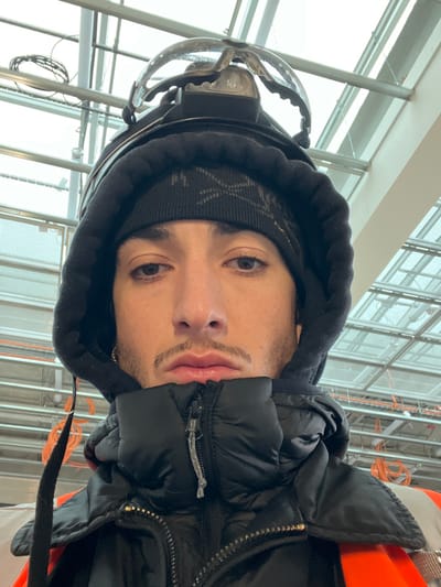 A close-up, head-and-shoulders portrait of a male subject is depicted in an indoor setting within Aulnay-sous-Bois, France. The subject, positioned centrally and looking directly at the camera, has a light mustache and a soul patch. He is wearing a black knitted beanie with a subtle pattern, a black hooded sweatshirt with the hood raised, and a black puffer jacket. An orange high-visibility garment is visible beneath the puffer jacket. A headlamp with a clear plastic visor is affixed atop the beanie. The background features a structural ceiling composed of metal beams and translucent or clear panels, indicative of a large commercial or industrial facility. Coiled orange cables are visible in the upper left background, attached to the ceiling structure.