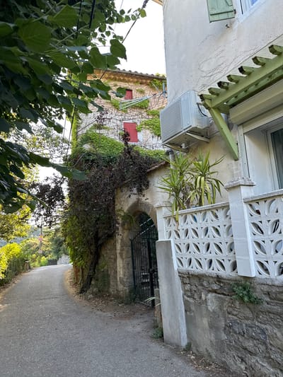 A narrow paved road curves uphill to the left, bordered by a stone wall and lush vegetation. On the right, a stone wall transitions to a lighter stucco wall adorned with ivy and an air conditioning unit. Further along the right, a decorative white concrete balustrade runs parallel to the road. A wrought iron gate is visible within an arched stone entryway in the background. The scene suggests a residential area in the French countryside, possibly Générargues.