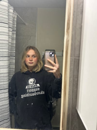 A mirror selfie shows a person with short blonde hair, wearing a black hoodie. The hoodie has a white graphic design of a skull with text printed around it. The person is holding a tan-colored iPhone in front of their face to take the picture. The background includes a shower curtain with gray and white stripes, tiled walls, and a portion of a white ceiling. The mirror has a light brown frame.
