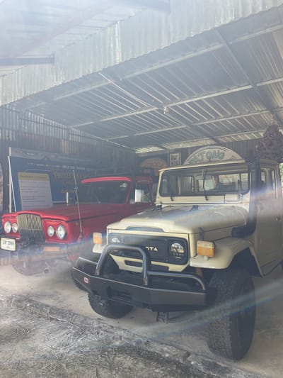 The image displays an interior view of a garage or shed, likely a museum or display area, located in Ban Yid, Thailand. Two classic off-road vehicles are parked on a concrete floor beneath a corrugated metal roof structure.

In the foreground, slightly to the right of the center, is a light beige (or cream-colored) Toyota Land Cruiser, identifiable as a J40 series. It features a heavy-duty black ARB branded front bumper and a black snorkel rising along the passenger side of the windshield. The front grille displays "TOYOTA" lettering, and the vehicle is equipped with modern LED headlights and amber turn signals. Knobby off-road tires are visible on the front right wheel.

To the left and slightly behind the Toyota is a red classic American SUV or truck, possibly a Jeep Gladiator or Wagoneer, distinguished by its prominent vertical slatted chrome grille and four circular headlights. A Thai license plate, "บห 2999" (BH 2999), is affixed to its front bumper.

The background features large banners and signs. Behind the red vehicle, a blue and white banner is visible with text including "AMAZING EXPERIENCE RIDE TOGETHER," "WORLD RIDE TOGETHER," and "PHUKETBIKE WE" (likely part of "PHUKET BIKE WEEK"). Below this, "วัตถุประสงค์ / Objective" is printed in Thai and English, followed by additional descriptive text. Above the beige Toyota, a curved sign with green lettering spells "PATONG." Other vintage-style signs, one mentioning "MOTORCYCLE," are partially visible further in the background.

The roof is constructed from corrugated metal sheets supported by visible metal trusses and beams. The concrete floor shows signs of wear, with a distinct raised concrete curb running horizontally across the lower part of the frame. The scene is brightly lit, with significant lens flare originating from the upper right quadrant of the image, indicating a strong, possibly natural, light source. No people are visible in the frame.