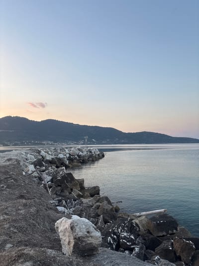 The image shows a rocky breakwater extending into a calm body of water, likely the sea, in Skala Potamias, Greece. The sky is a pale, clear blue with a hint of pink and orange near the horizon, suggesting it is either dawn or dusk. The water is a serene shade of blue-green, with gentle ripples.

On the left side of the frame, a mound of dark, coarse material, possibly sand mixed with seaweed or debris, is piled up against the rocks. The breakwater itself is constructed of large, irregularly shaped grey and white rocks. A weathered, pale piece of driftwood rests on some of the rocks near the water's edge. In the distance, a low, forested mountain range forms the backdrop, with a small settlement visible along its base. A slender white pole with a light or marker at its top stands on the breakwater, further out towards the water. The scene is peaceful and devoid of human activity.