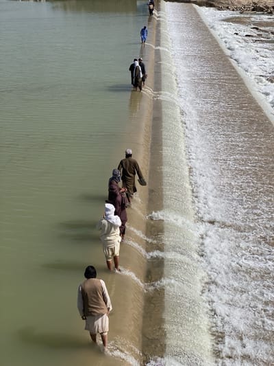 The image depicts a group of people walking across a concrete dam or weir in a river or large stream. The water appears murky and light brown in color.

People:
*   There are at least nine individuals visible.
*   They are dressed in traditional Afghan clothing, including long shirts, vests, and head coverings (turbans or skullcaps).
*   Most are walking barefoot or with their feet in the water that is flowing over the dam.

Objects:
*   A long concrete dam or weir spans the width of the waterway.
*   Water is flowing over the edge of the structure.
*   The far bank appears to be sandy or rocky.

Actions:
*   The people are walking across the dam in a single line.

Environment:
*   The scene is outdoors in daylight.
*   The landscape appears arid or semi-arid.
*   The location is unspecified, but the prompt suggests it may be near Kim, Afghanistan.