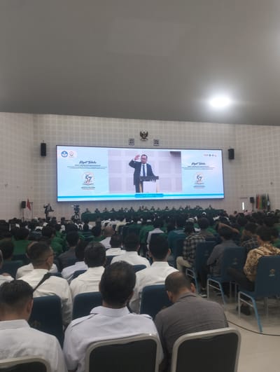 This is a full shot image of a large indoor gathering, likely a university event, taken from a low angle. The scene is dominated by a large digital screen at the front of a spacious auditorium, displaying an image of a man in a suit speaking at a podium. The screen also features text and logos in Indonesian, indicating the event is titled "Rapat Tahunan" (Annual Meeting) and is related to "SENAT UNIVERSITAS MALIKUSSALEH" (Malikussaleh University Senate). The date "22 Juni 2023" is visible on the screen.
In front of the screen, a speaker stands at a podium, addressing the audience. To the left of the podium, a red flag on a stand is visible. A camera on a tripod is positioned in the center, facing the stage.
The audience is seated in rows of chairs, filling a significant portion of the auditorium. Many attendees are wearing white shirts, while others are in green shirts or casual attire. Their backs are mostly to the camera, as they are facing the stage and screen.
The environment appears to be a modern, well-lit auditorium with white tiled walls and a high ceiling. The lighting fixtures are visible on the ceiling, and a bright light source is evident. Flags of various colors are arranged on stands to the right side of the stage area. The overall impression is of a formal academic or public assembly. The specific location in Reuleut Timu, Indonesia, is inferred from the context of "UNIVERSITAS MALIKUSSALEH", which is a university located in that region.