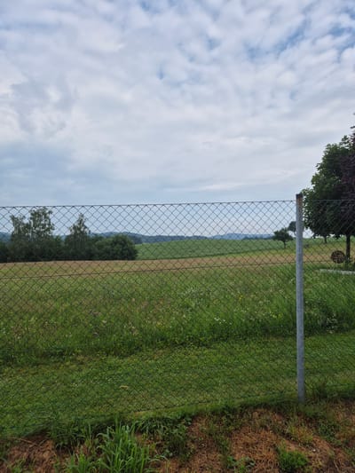 The image shows a chain-link fence in the foreground, with a natural landscape beyond. The fence is green and appears to be made of metal. Behind the fence, there is a field with green grass and some wildflowers. Further in the distance, there are rolling hills covered in trees and fields. The sky is cloudy with patches of blue sky visible. On the right side of the image, there are trees, one with dark red leaves, and a metal pole. The scene is outdoors and appears to be in a rural or semi-rural area. There are no people visible in the image. The location is specified as Au, Austria.