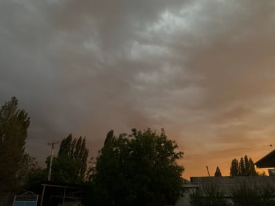 The image depicts an overcast sky with a warm, orange glow on the right side, suggesting either sunrise or sunset. Below the sky, a line of tall, slender trees stands on the left, interspersed with a wider, leafy tree in the center. In the foreground, there are parts of buildings with corrugated metal roofs, a covered structure with a flat roof, and a blue gate with white bars. A utility pole with wires is visible among the trees. The scene appears to be a rural or semi-urban residential area. The weather seems unsettled, with thick, dark clouds dominating the upper portion of the sky. The overall mood is somewhat dramatic due to the contrast between the dark clouds and the warm light. There is no visible text in the image. The location is likely Myrza-Ake, Kyrgyzstan.
