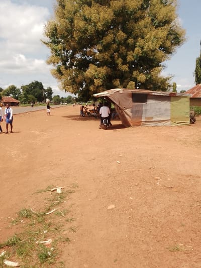 The image depicts a rural roadside scene in Adikpo, Nigeria. The ground is unpaved and reddish-brown. A small, makeshift stall constructed from corrugated metal and fabric is situated on the right side of the frame. A person wearing a white t-shirt is seated on a motorcycle parked in front of the stall. To the left, two young women in school uniforms, a white blouse and a blue dress, are walking. In the background, there are several buildings with corrugated metal roofs, trees, and people walking on a paved road. The sky is partly cloudy.