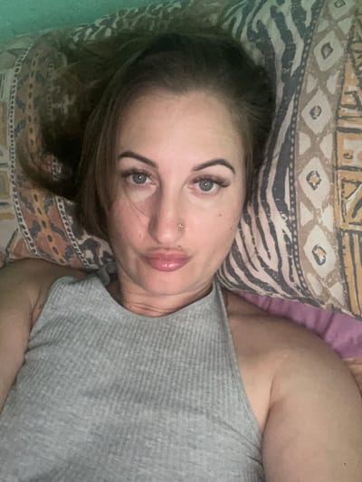 A close-up, overhead selfie shows a woman lying on a bed with a patterned pillow. She has brown hair and light blue eyes, and she is wearing a grey ribbed tank top. Her lips are pursed, and she has a nose ring. The pillow behind her features a repeating pattern of geometric shapes and stripes. The background bedding is a mix of green and purple. The image appears to be taken indoors, in a domestic setting. There is no indication of the woman's location in Zarichne, Ukraine.