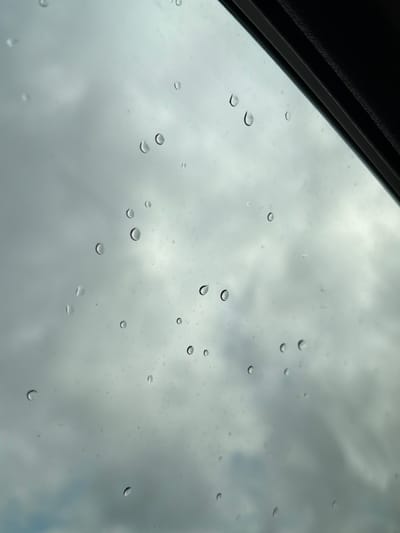 The image depicts a close-up, slightly angled view of raindrops on a glass surface, likely a window or windshield. The background is a hazy, overcast sky with diffused light, suggesting cloudy weather. The raindrops vary in size and shape, with some appearing as perfect spheres, while others are elongated or distorted. The glass surface itself seems to be the primary subject, with the raindrops creating a pattern across it. The top right corner of the image shows a dark, textured edge, possibly the frame of the window or a car's sunroof. There are no people or any other objects visible in the image. The scene appears to be an everyday observation of weather, focusing on the aesthetic of water droplets on glass. The specific location in Oreshniki, Russia is not discernible from the visual information provided in the image.