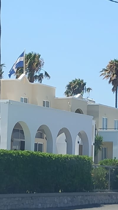 The image shows a building with a Greek flag flying from a flagpole. The building appears to be in a sunny, outdoor environment. The architecture of the building is characterized by arches and a light-colored facade. Palm trees are visible around the building, suggesting a warm climate. The image is taken from a slightly low angle, looking up at the building and the flag. The sky is clear and blue. There is a green hedge in the foreground. The scene suggests a location in Greece, possibly near the sea given the palm trees.