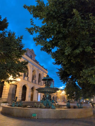 The image depicts the Ganja Philharmonic Hall in Ganja, Azerbaijan, at dusk. A prominent bronze fountain, adorned with sculpted figures, sits in the foreground. Behind the fountain, the neoclassical facade of the Philharmonic Hall is illuminated by warm artificial lights, highlighting its arched entrances and classical architectural details. Lush green trees with large leaves frame the scene, their branches reaching towards the twilight sky, which transitions from a deep blue to a lighter hue near the horizon. The street in front of the building appears to be paved. In the background, more trees and some distant lights suggest the presence of an urban environment. A few indistinct figures can be seen walking in the distance. The overall atmosphere is serene and elegant.
