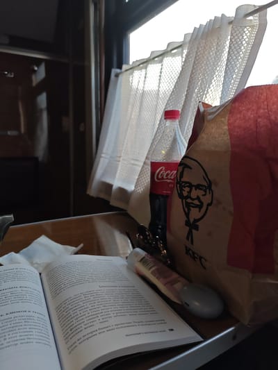 The image depicts a scene inside a train carriage, with a window covered by white curtains in the background. In the foreground, an open book rests on a surface alongside a half-empty Coca-Cola bottle, a KFC bag, and a tube of lotion. The text in the book is in Russian. The location, inferred from the text in the book, is likely Мажэйкаўскі сельскі Савет, Belarus.