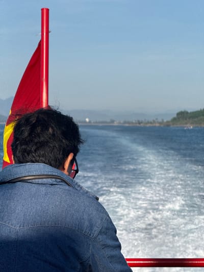 In An Hải, Vietnam, a person wearing a denim jacket looks out at the wake of a boat. The person has dark, curly hair and is wearing glasses. To the left, a red pole with a red and yellow flag is visible. The water is blue and choppy, with a clear trail of white foam stretching behind the boat. In the distance, a hazy shoreline with greenery and a few buildings can be seen under a clear blue sky. The image is captured from a high angle, looking down and behind the person.