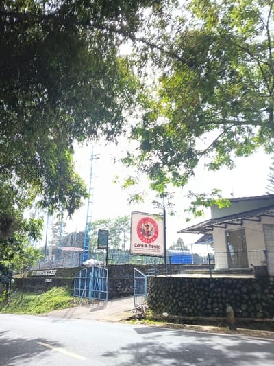 The image captures an outdoor scene in Carcar, Philippines, presenting a roadside view of a sports and dining facility on a bright, sunny day. The asphalt road in the foreground is dappled with shadows, indicative of clear weather and strong sunlight, likely morning or afternoon.
The upper part of the frame is filled with the dense, green foliage of large trees, which provide natural shade and frame the view of the facility. A low stone wall forms a boundary between the road and the property, with two blue metal gates, partially open, providing an entrance.
Behind the gates and wall, the facility extends. On the left, a sign confirms the presence of a "FUTSAL SOCCER" area. Tall, blue stadium light poles are visible in the background, suggesting a sports field enclosed by blue fencing or netting.
A prominent, large rectangular sign on the right advertises "THE OLD BRICK CAFE & RESTO." The sign features a logo with a stylized character, possibly a chef, and includes additional text: "Love Music • Good Food • Healthy." Below these, further details are visible, though less clear, seemingly listing menu items like "Pizza, Burger, Pasta, Coffee," and "Ice Cream." Adjacent to this sign is a building, likely the cafe and resto, characterized by a dark-tiled roof and light-colored walls.
No individuals are visible in the scene, and no specific activity or event is actively unfolding. The image conveys a neutral, pleasant atmosphere, highlighting the commercial and recreational aspects of the location.